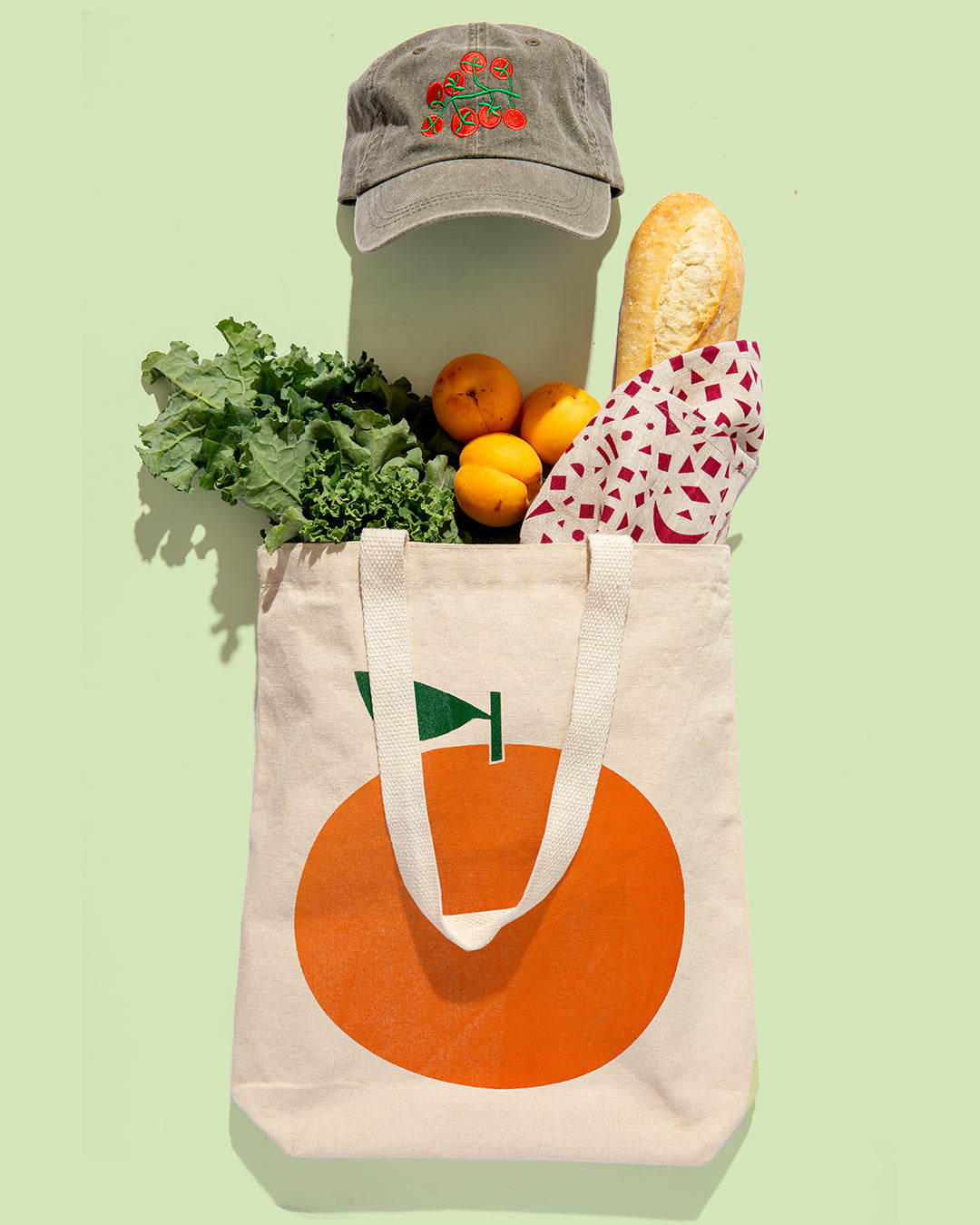 A cream canvas tote bag with a large orange graphic and green flag detail, filled with kale, oranges, a baguette, and a patterned cloth, with a gray embroidered baseball cap resting above it on a pale green background.