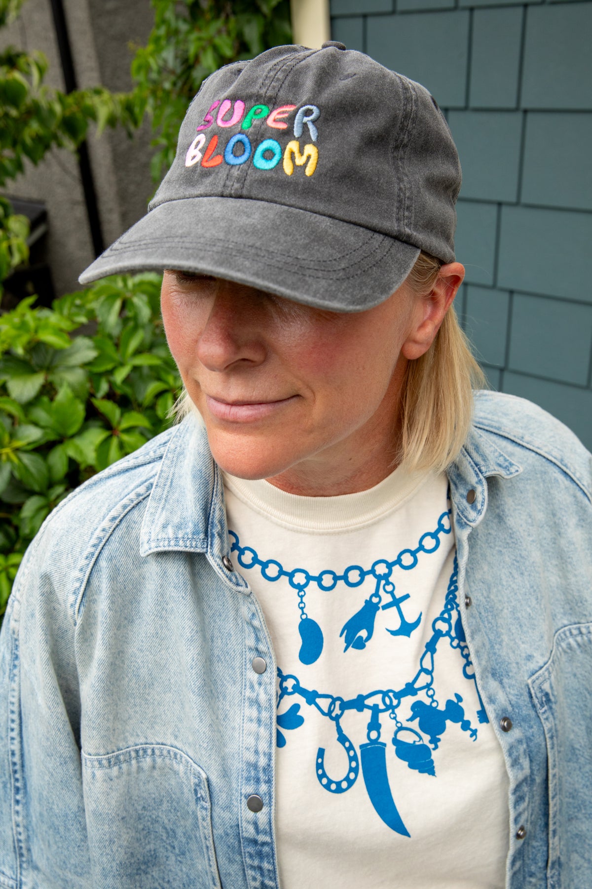 A woman wearing a dark gray baseball cap with 'SUPER BLOOM' embroidered in colorful letters, styled with a light blue denim shirt and a white graphic t-shirt.