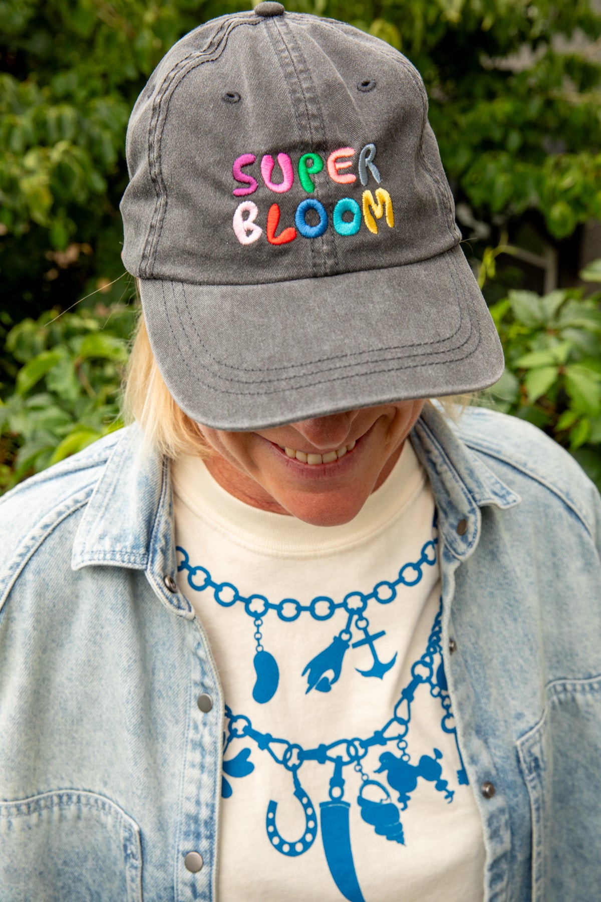 A person outdoors wearing a gray baseball cap with colorful embroidered text reading 'SUPER BLOOM' across the front, layered with a light blue denim jacket over a cream graphic T-shirt.