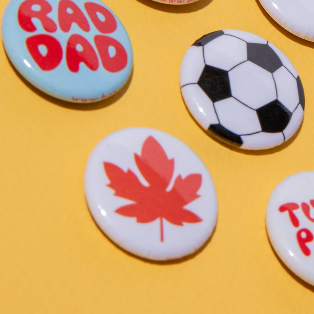 A close-up of several round pin-back buttons on a yellow background. One button is light blue with red text reading 'RAD DAD,' another is a black-and-white soccer ball, and a third is white with a red maple leaf.