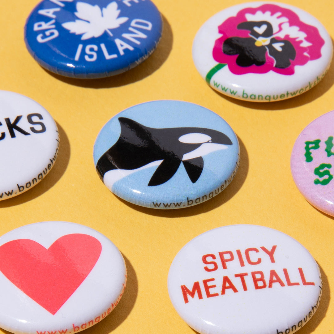 A group of round white button badges with colorful printed designs, including a blue Canada Islands badge, a pink flower, an orca, a red heart, and a red 'Spicy Meatball' text design, arranged on a yellow background.