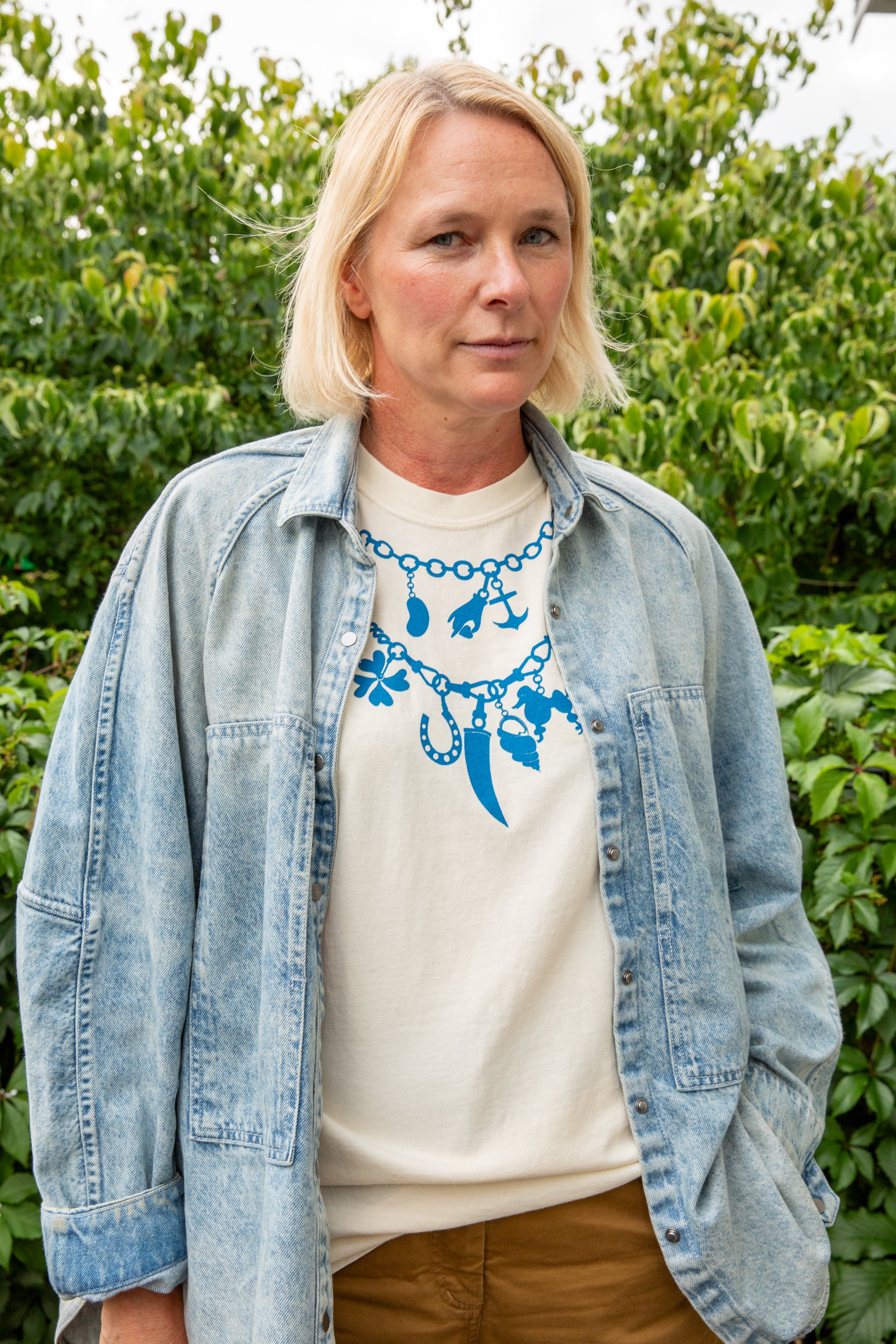 A woman with blonde hair stands outdoors in front of green foliage, wearing a light blue denim shirt over a cream t-shirt with a blue charm necklace graphic, and brown pants.
