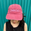 Pink text in bubble letters that says SNACKS on a raspberry pink hat work by a woman