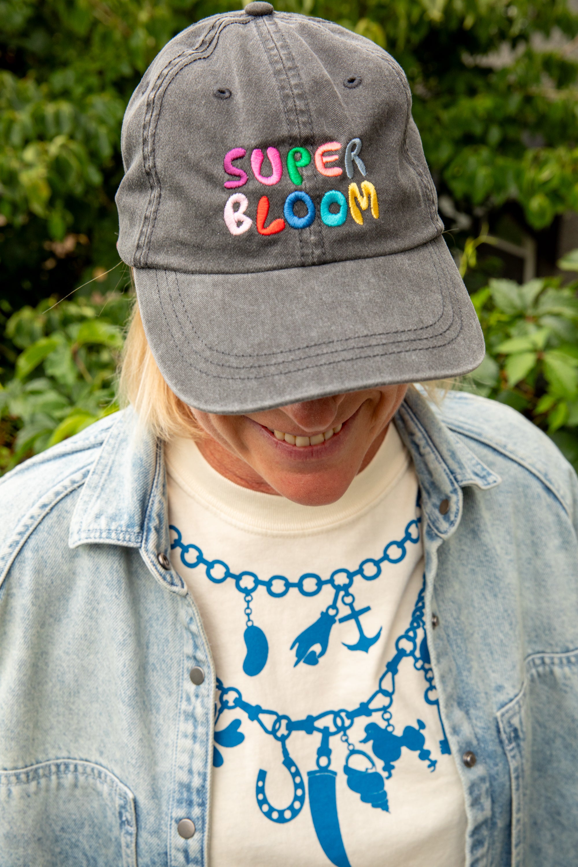 A person outdoors wearing a gray baseball cap with colorful embroidered text reading 'SUPER BLOOM' across the front, layered with a light blue denim jacket over a cream graphic T-shirt.