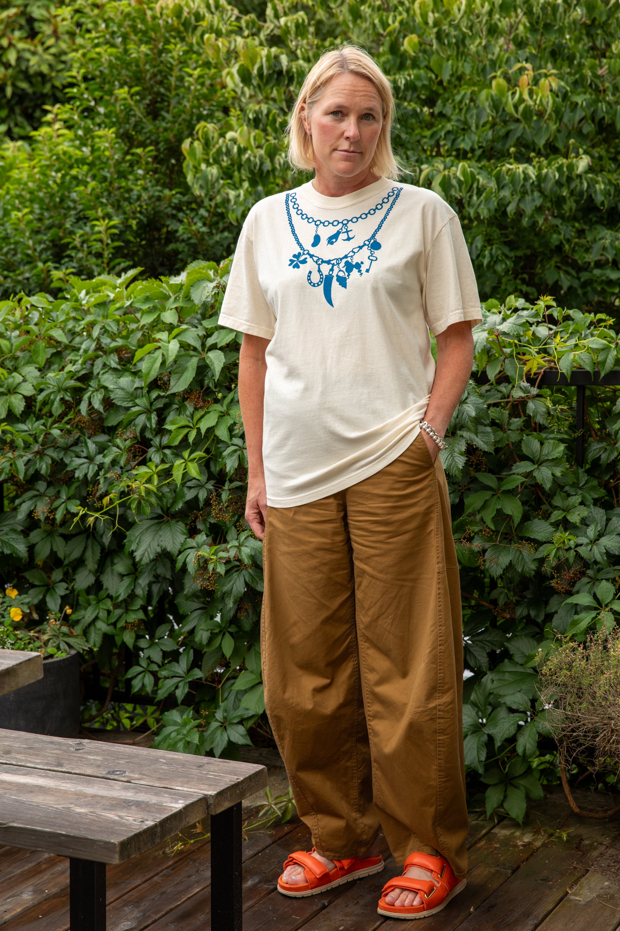 A woman standing outdoors in front of green foliage, wearing a cream-colored graphic t-shirt with a blue charm necklace print, loose brown pants, and orange sandals.