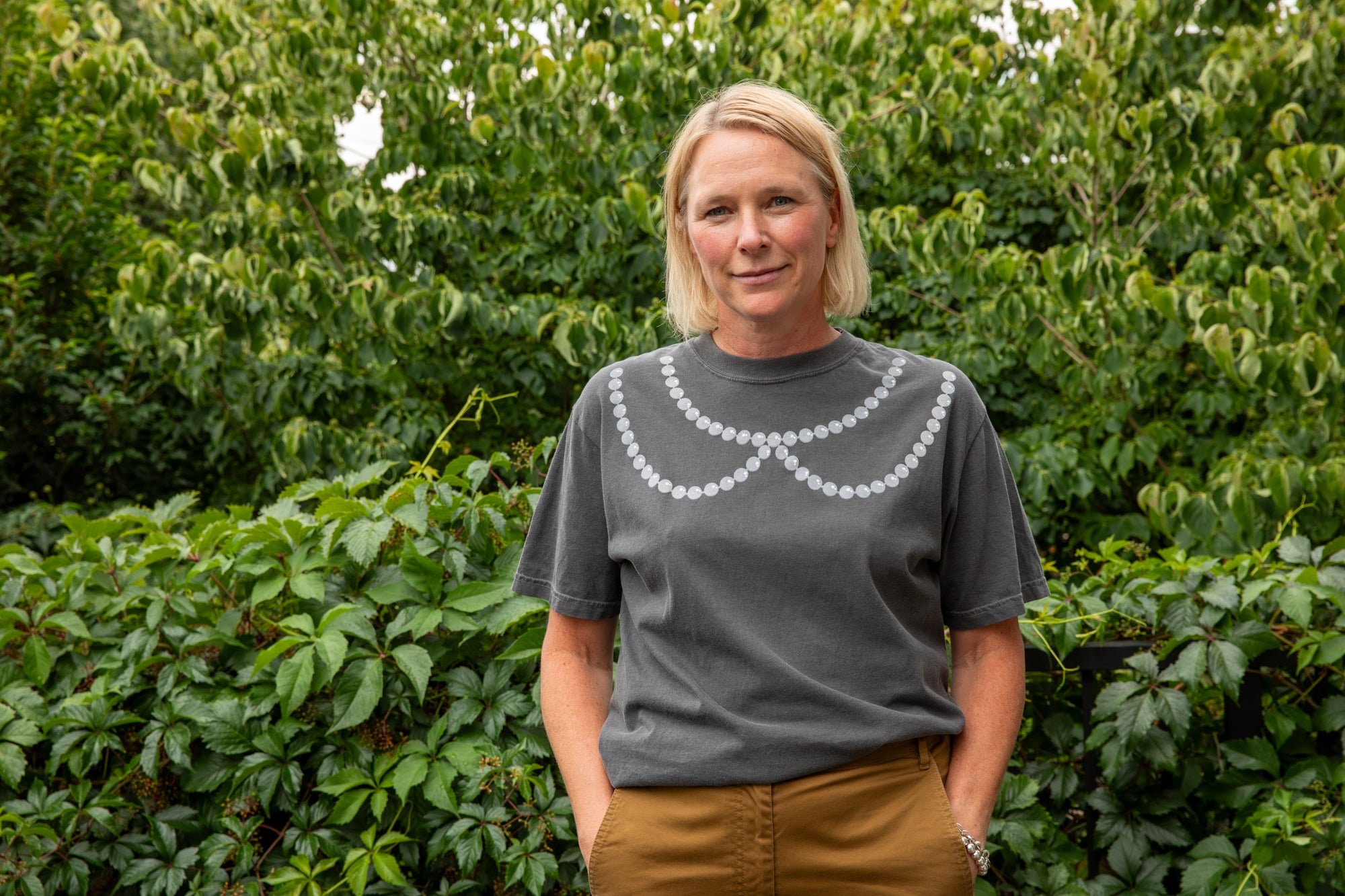 A woman with blonde hair stands outdoors in front of a green hedge, wearing a dark gray t-shirt with a white scalloped collar design and brown pants.