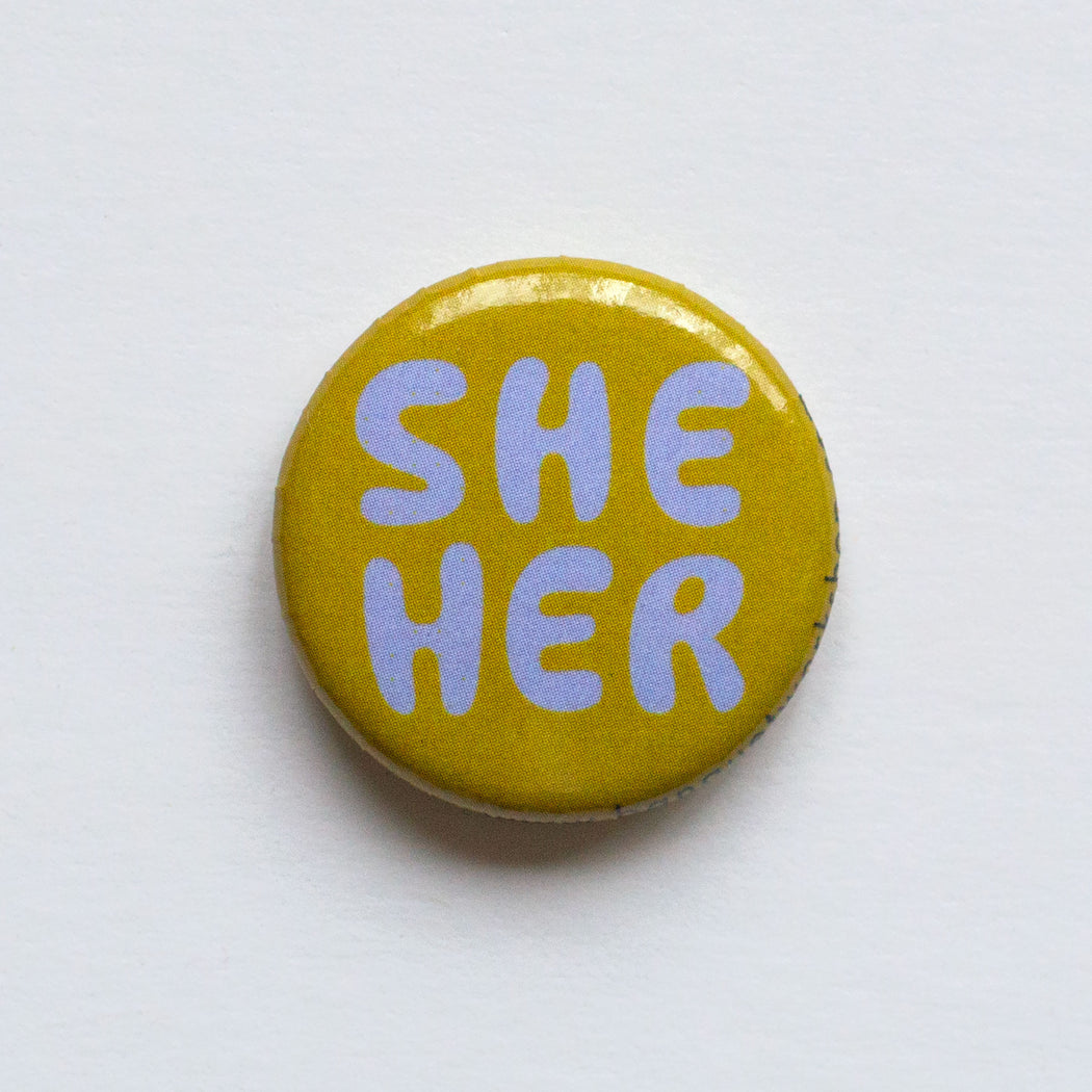 one inch lapel button reads "She Her" in funny lilac lettering on mustard