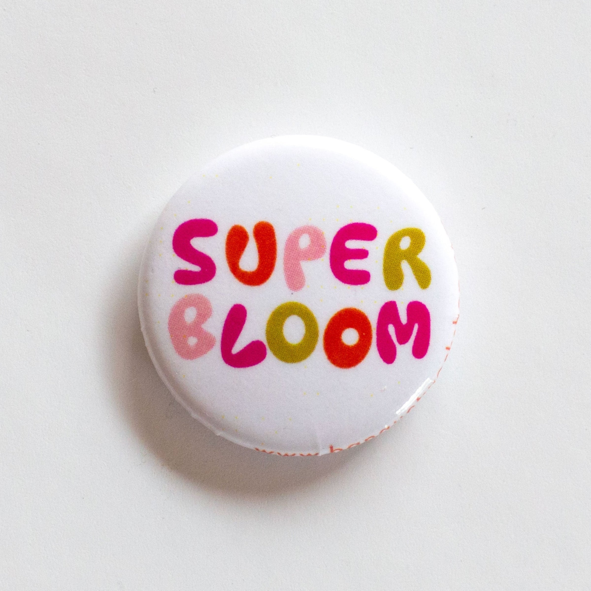 one inch button says "super bloom" in multicoloured lettering